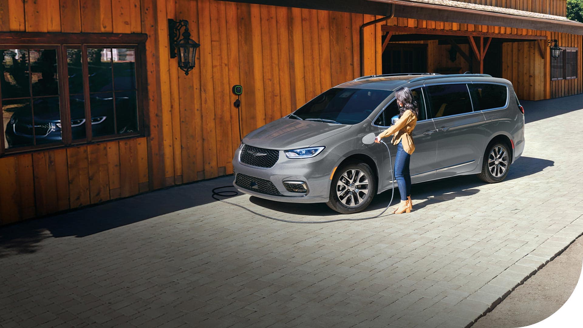 A driver-side front angle of a silver 2026 Chrysler Pacifica Plug-In Hybrid Pinnacle parked at a farmers market that has a built-in charging station, with a woman plugging its charging cord into the minivan's charging port.