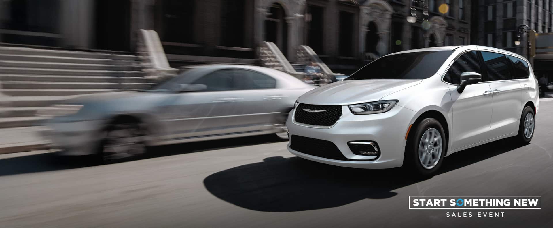 A white 2026 Chrysler Pacifica Select driving down a city street. Start Something New Sales Event.