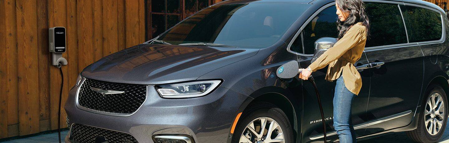 A woman beside a 2022 Chrysler Pacifica Pinnacle Hybrid plugging a charging cable into the vehicle's charging port.