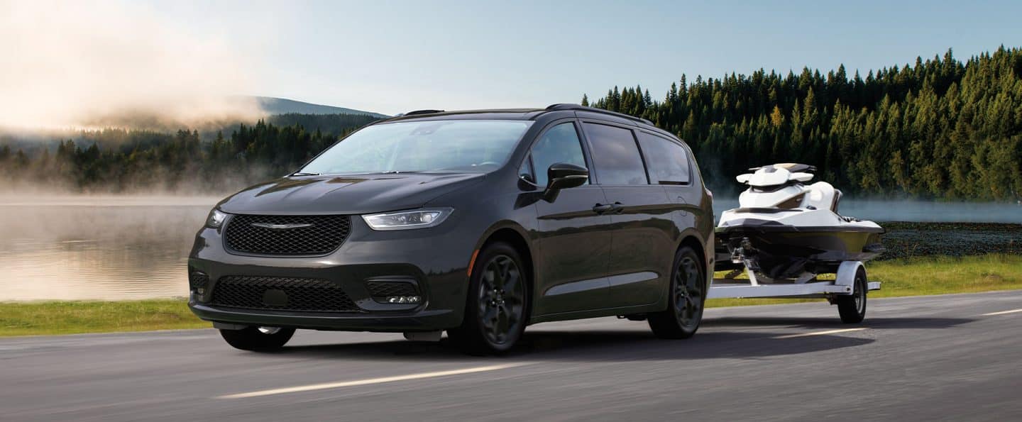 Una Chrysler Pacifica Limited 2025 negra circulando por una carretera en frente de un lago, acarreando una moto de agua.