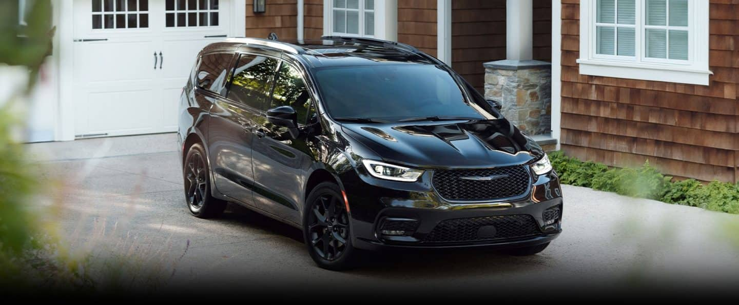 Ángulo delantero del lado del pasajero de una Chrysler Pacifica Limited 2025 negra, estacionada en la entrada de garaje de una casa moderna.