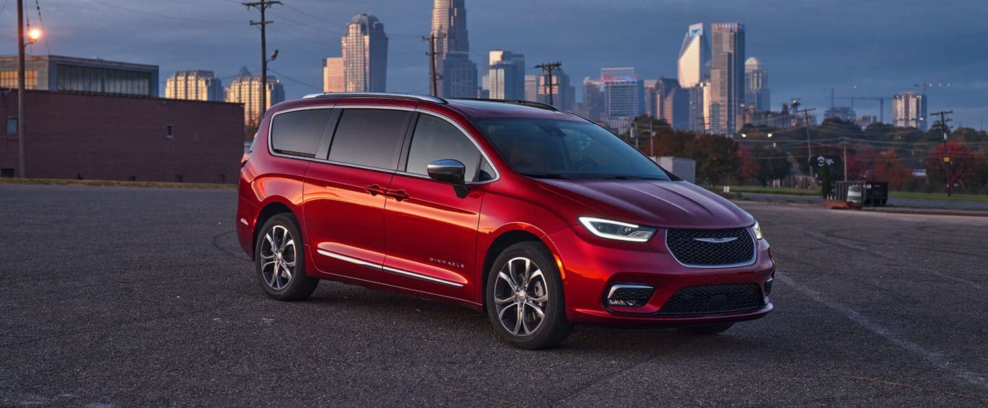 Ángulo delantero del lado del pasajero de una Chrysler Pacifica Pinnacle 2025 roja estacionada en un lote al atardecer con edificios altos de fondo.