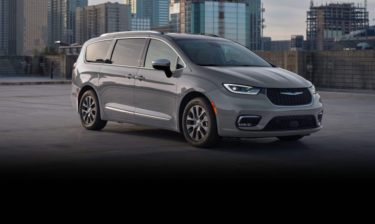 Ángulo delantero del lado del pasajero de una Chrysler Pacifica Pinnacle Plug-in Hybrid 2025 gris estacionada en el techo de una estructura de estacionamiento, con edificios altos de fondo.