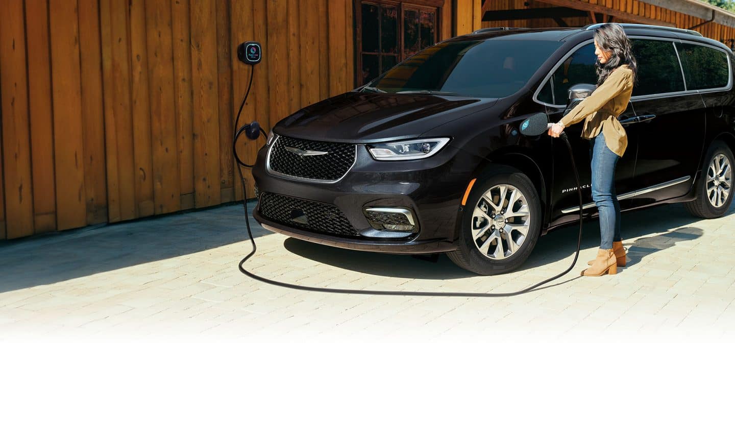 Ángulo delantero del lado del conductor de una Chrysler Pacifica Pinnacle Hybrid 2025 enchufable de color negro, estacionada fuera de un edificio con una estación de carga. Una mujer enchufando el cable de carga de la estación en el puerto de carga del vehículo.