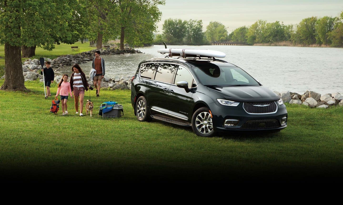 Una Chrysler Pacifica Pinnacle Hybrid 2025 enchufable de color negro, estacionada a la orilla de un lago con una tabla de pádel atada al portaequipaje y una familia de cuatro con su perro, caminando hacia el vehículo. Evento de ventas de Black Friday. 