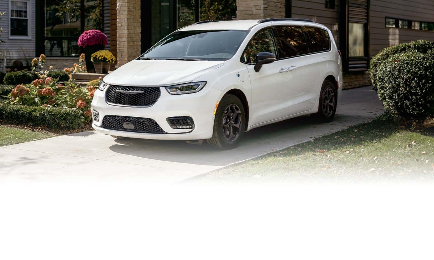 Ángulo delantero del lado del conductor de una Chrysler Pacifica Select Plug-in Hybrid 2025 blanca con el paquete apariencia Premium S, estacionada en la entrada de una casa en un vecindario.