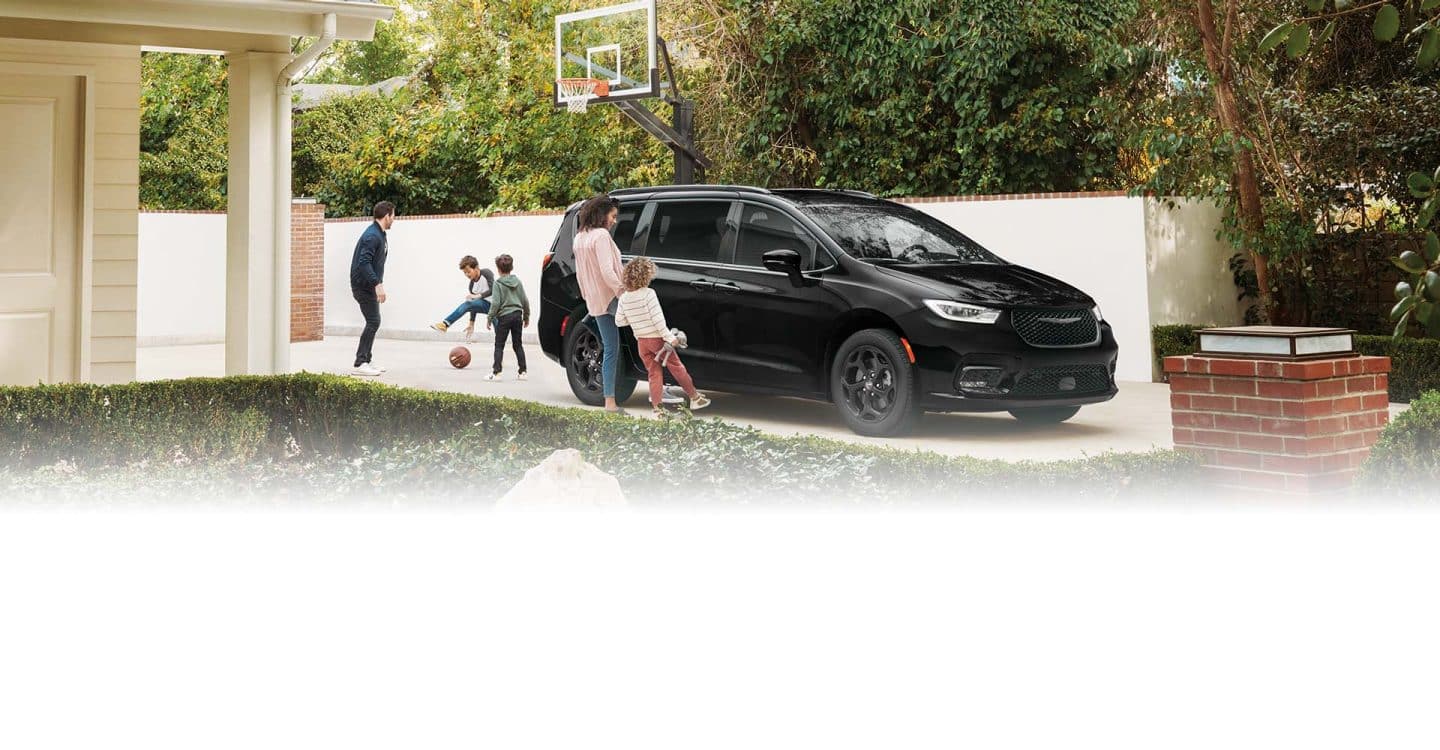 Una Chrysler Pacifica Select Hybrid 2025 enchufable con el paquete apariencia Premium S, estacionada en la entrada de una casa en un vecindario mientras un padre y sus dos hijos juegan al básquetbol. Una madre y su hija de pie junto a la puerta lateral corrediza eléctrica manos libres mientras cada una balancea un pie debajo de la puerta para abrirla.