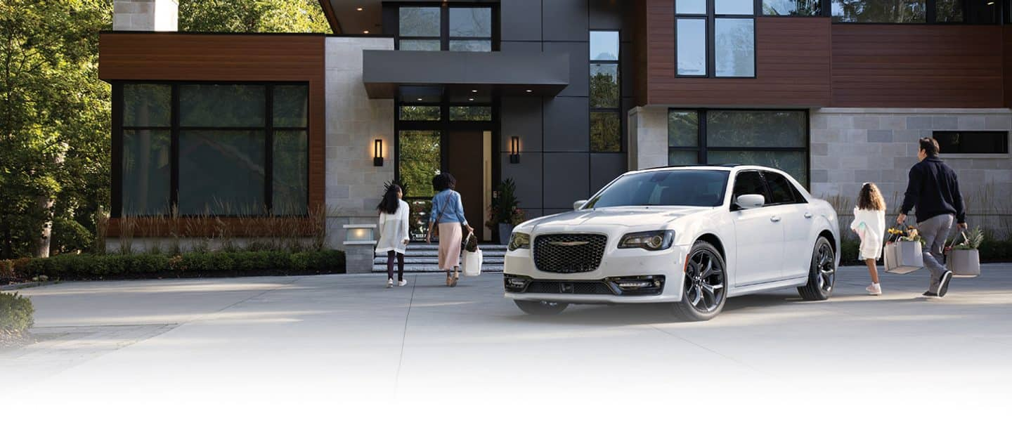 A white 2023 Chrysler 300 300S parked in the driveway of a large, contemporary home with a family of four walking from the vehicle to the front door.