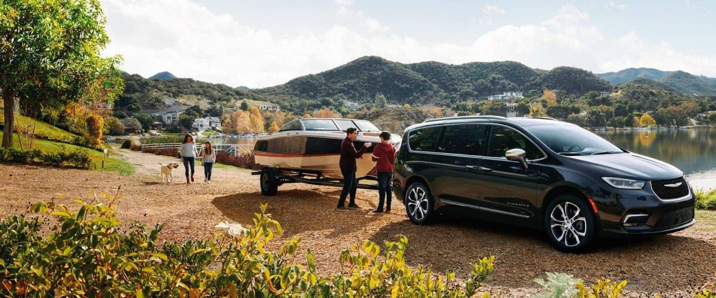 Un ángulo del lado del pasajero de una Chrysler Pacifica Pinnacle modelo 2024 de gasolina negra estacionada junto a un lago, remolcando una lancha a motor, con una familia de cuatro personas y su perro cerca. Concluya el evento de ventas del año.