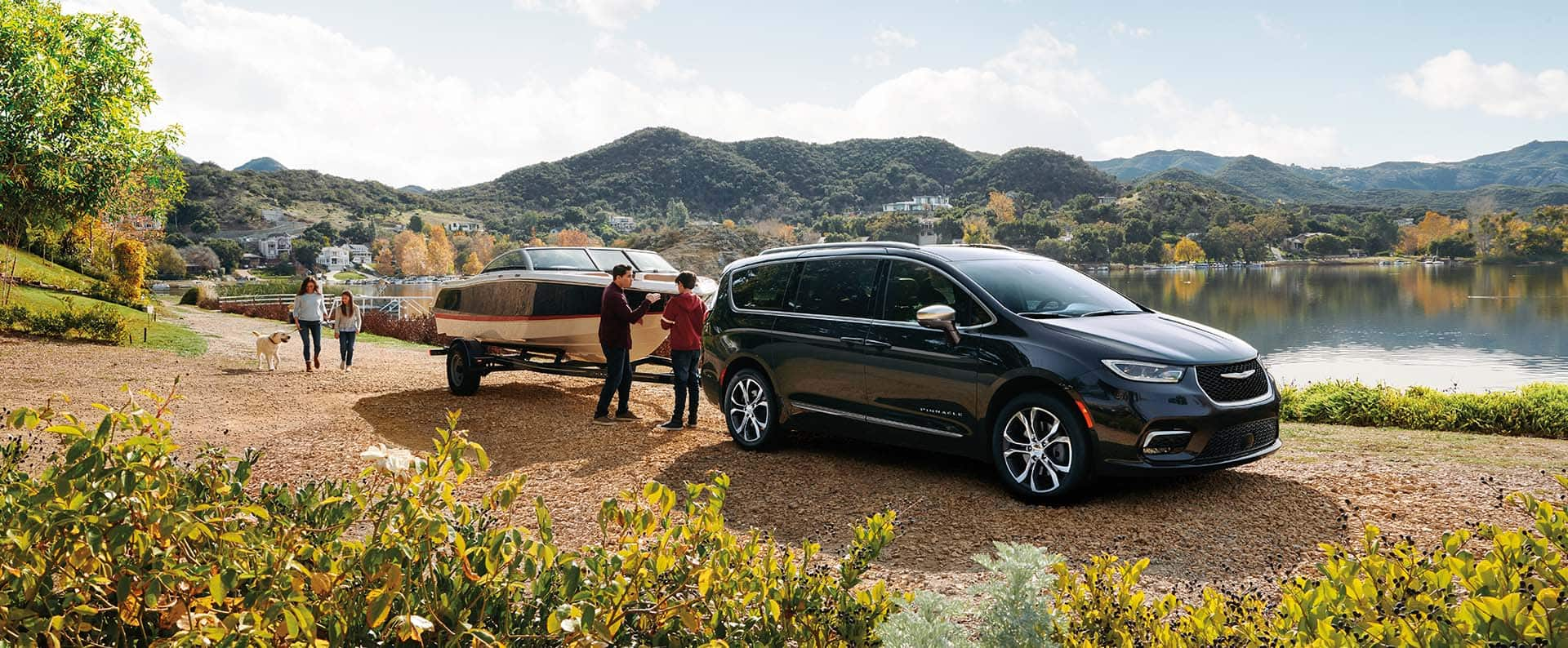 A black 2024 Chrysler Pacifica Pinnacle parked beside a lake, towing a motorboat, with a family of four nearby.