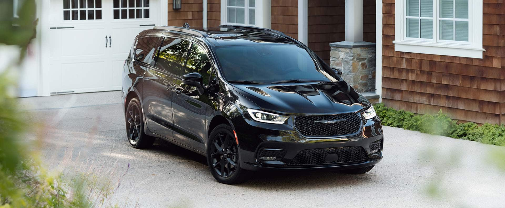 A passenger-side front angle of a black 2025 Chrysler Pacifica Limted parked in the driveway of a contemporary home.