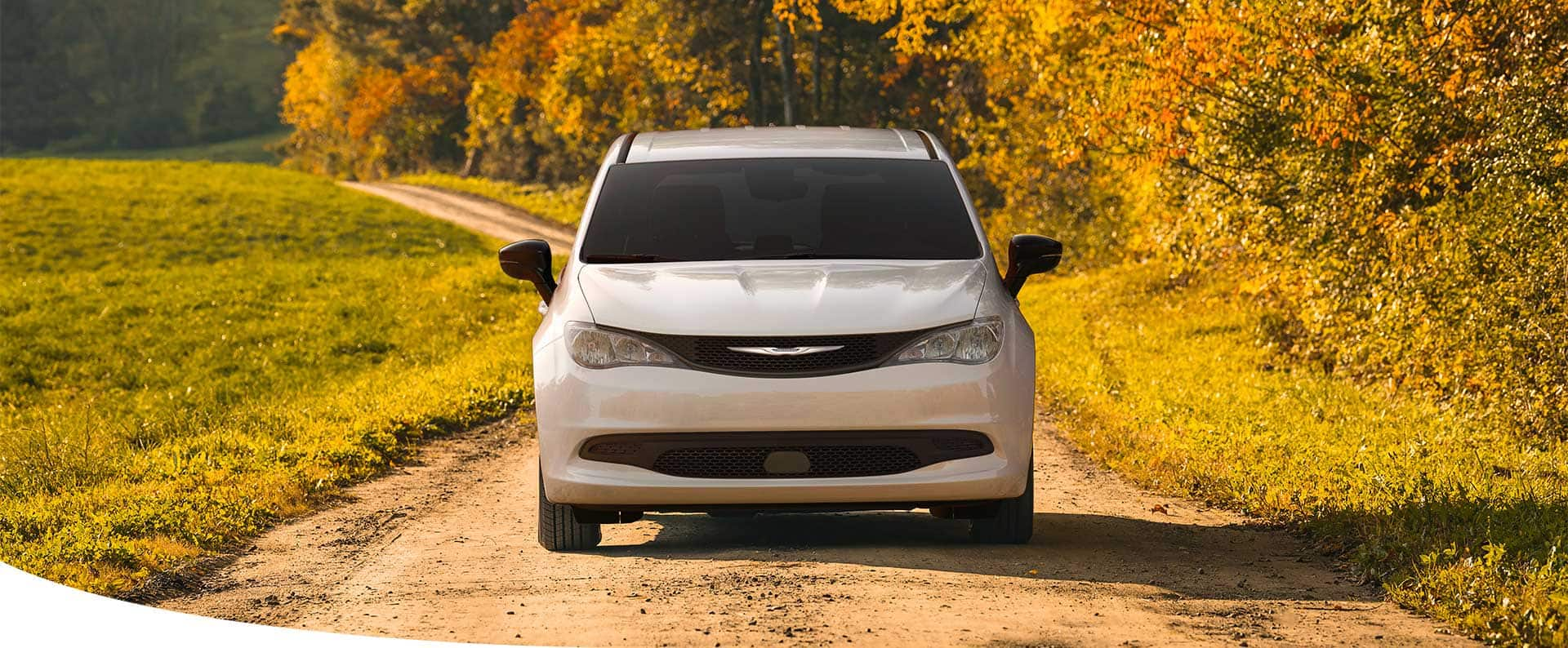 A head-on angle of a white 2025 Chrysler Voyager LX traveling on a dirt road in the country.