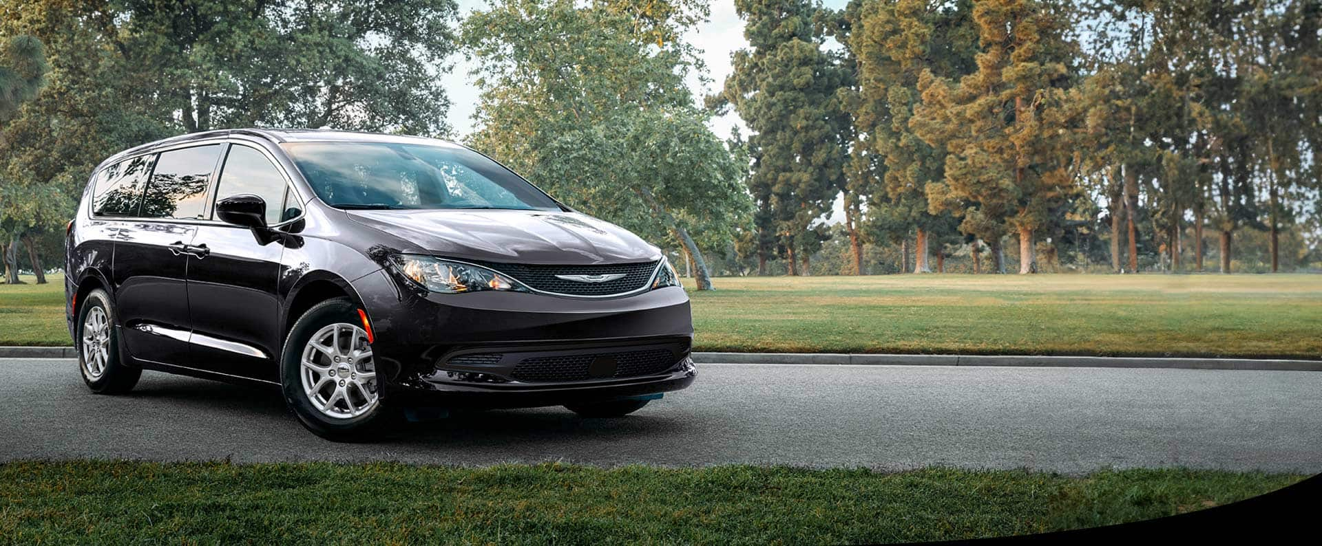 A front pasengers side view of a black 2025 Chrysler Voyager LX in a parking lot of a park.