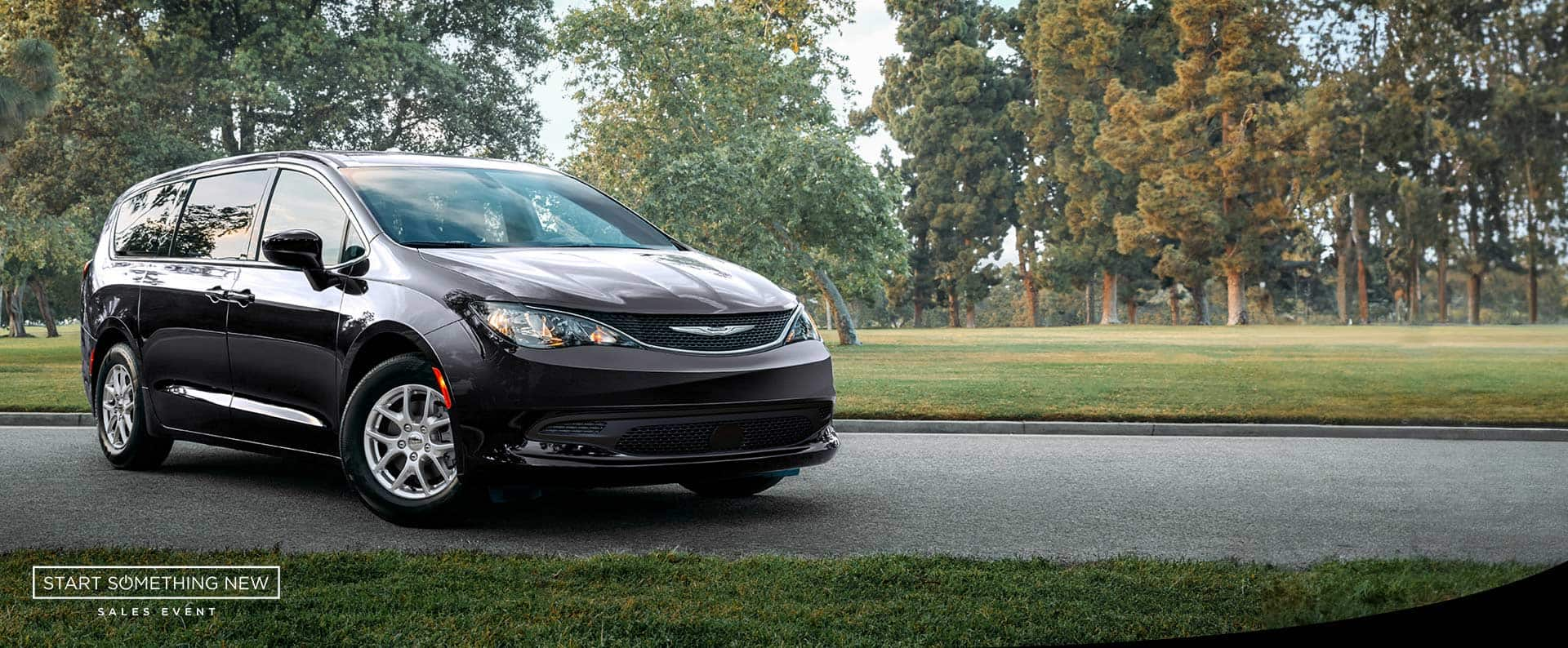 A front pasengers side view of a black 2025 Chrysler Voyager LX in a parking lot of a park. Start Something New sales event. 