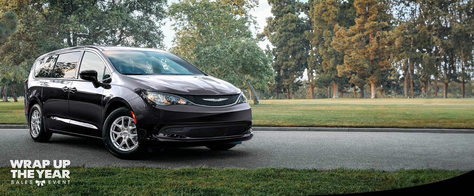 A front pasengers side view of a black 2025 Chrysler Voyager LX in a parking lot of a park. Wrap up the Year sales event.