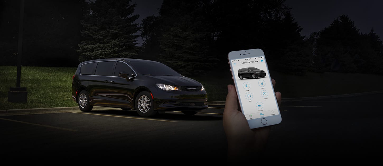 In the foreground a person holding a smartphone that is displaying the Chrysler app. In the background a black 2025 Chrysler Voyager LX. 