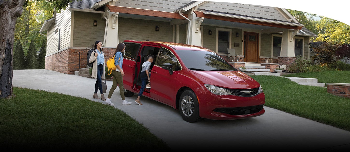 A red 2025 Chrysler Voyager LX parked in the driveway of a home with its passenger-side sliding door open and one of two children entering the vehicle, along with a woman.