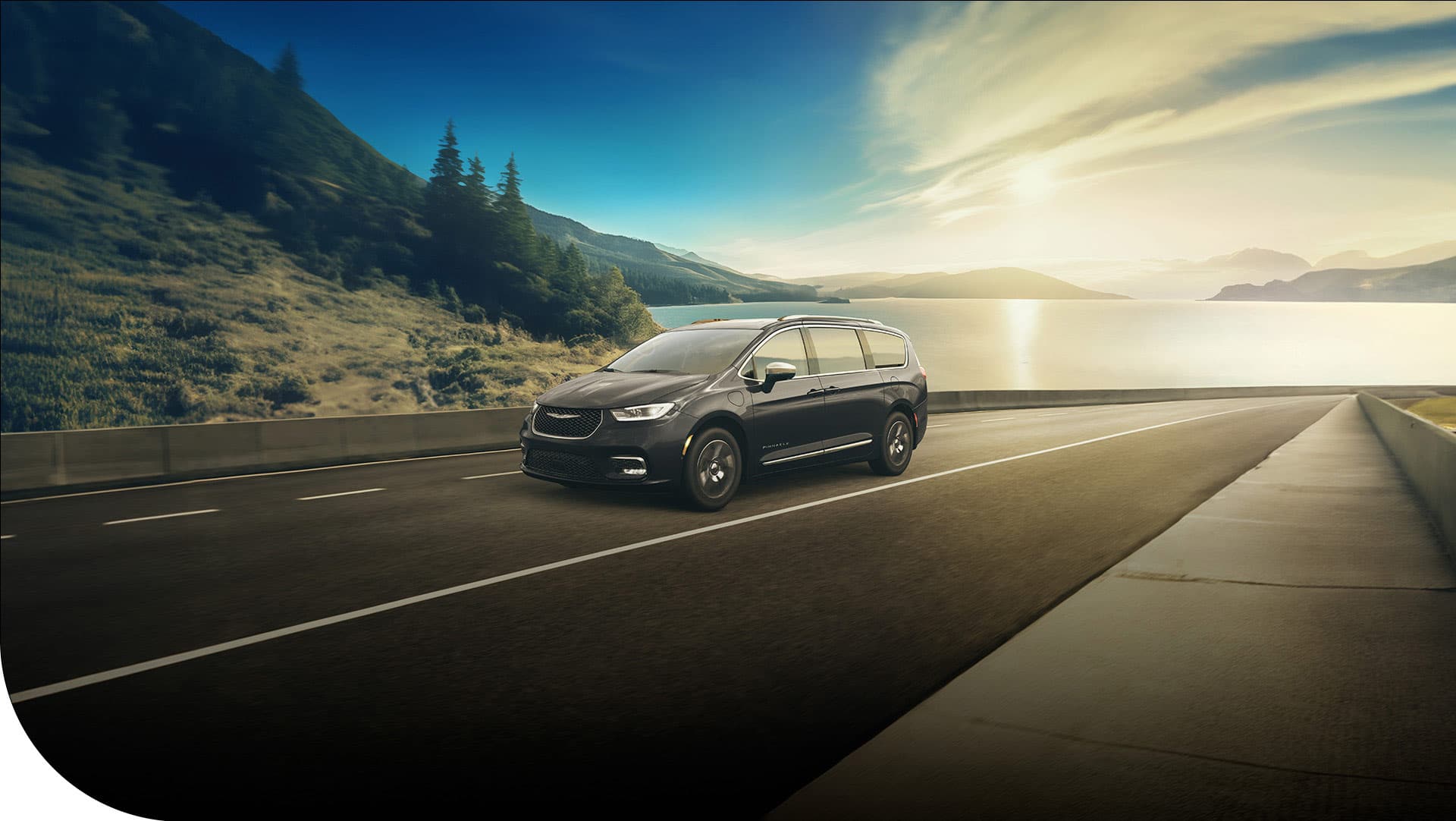 A black 2026 Chrysler Pacifica Plug-in Hybrid Pinnacle being driven on a highway beside a lake, with mountains in the background.
