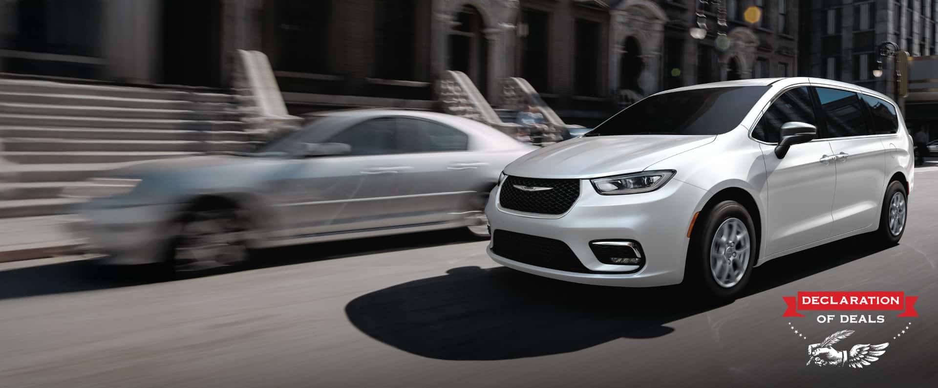 A white 2026 Chrysler Pacifica Select driving down a city street. Declaration of Deals Sales Event.