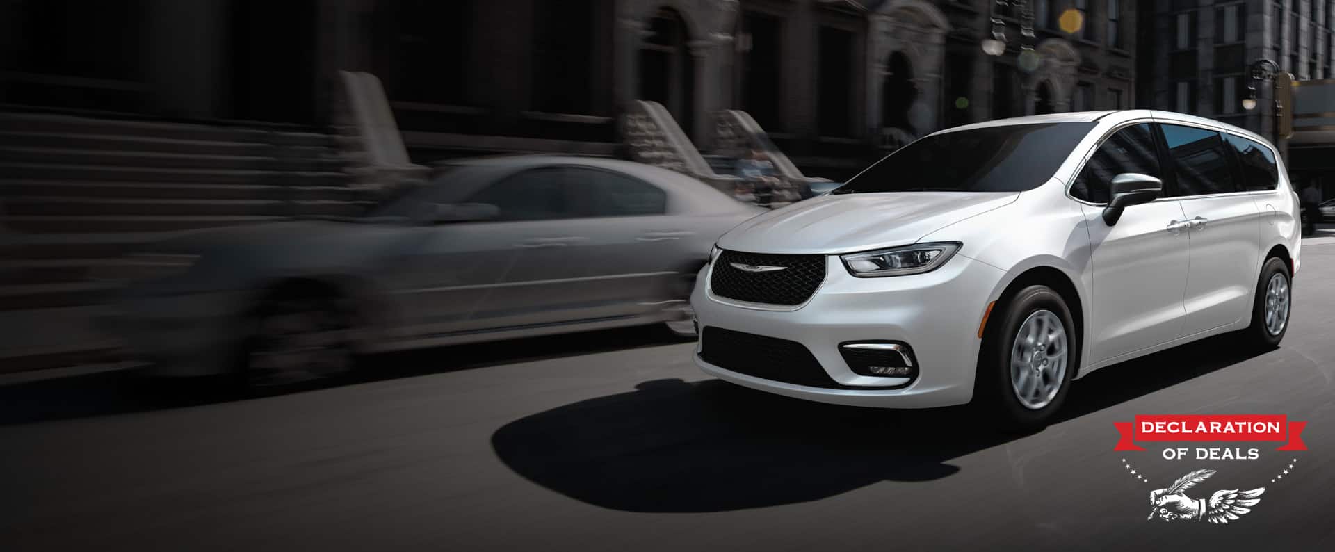 A white 2026 Chrysler Pacifica Select driving down a city street. Declaration of Deals Sales Event.