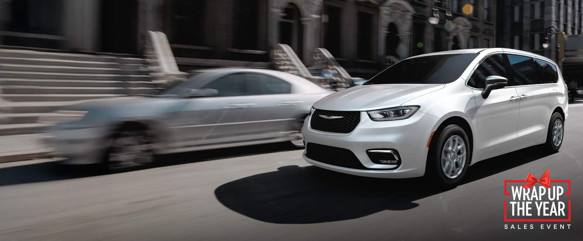 A white 2026 Chrysler Pacifica Select driving down a city street. Wrap up the Year sales event.
