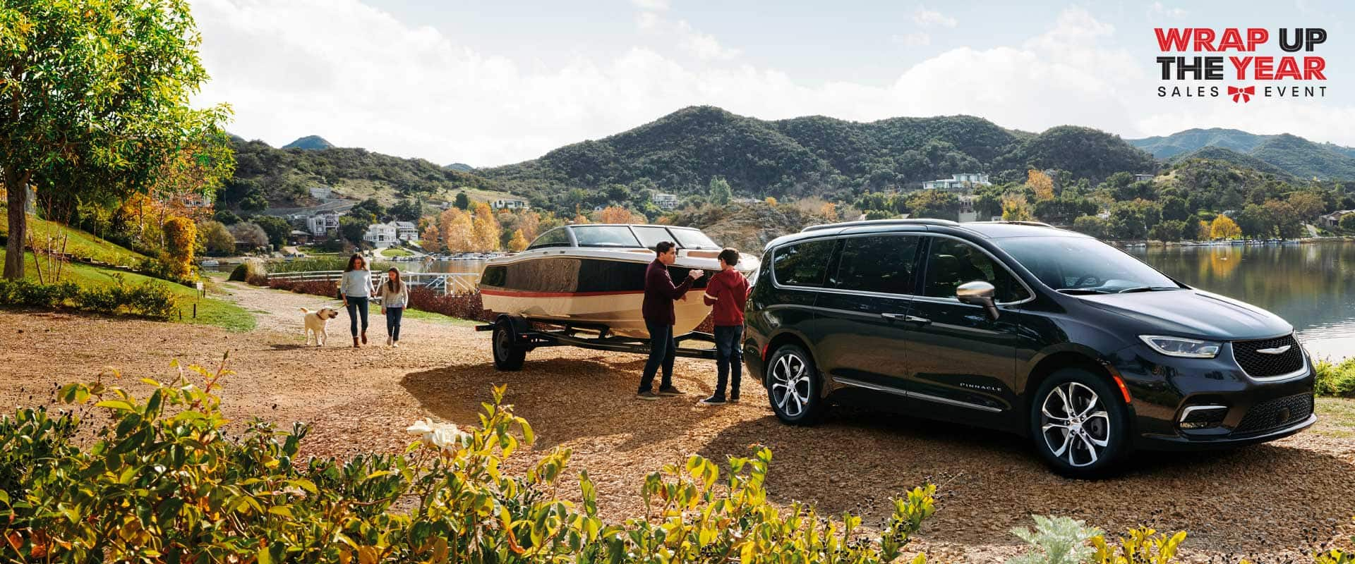 A passenger-side angle of a black 2024 Chrysler Pacifica Pinnacle gas model parked beside a lake, towing a motor boat, with a family of four and their dog nearby. Wrap up the Year sales event.